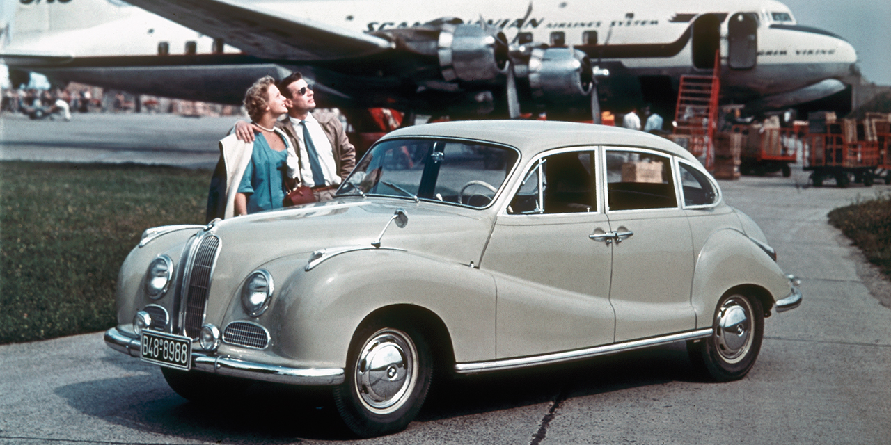 Paar hinter BMW 501 mit Flugzeug im Hintergrund (1951)