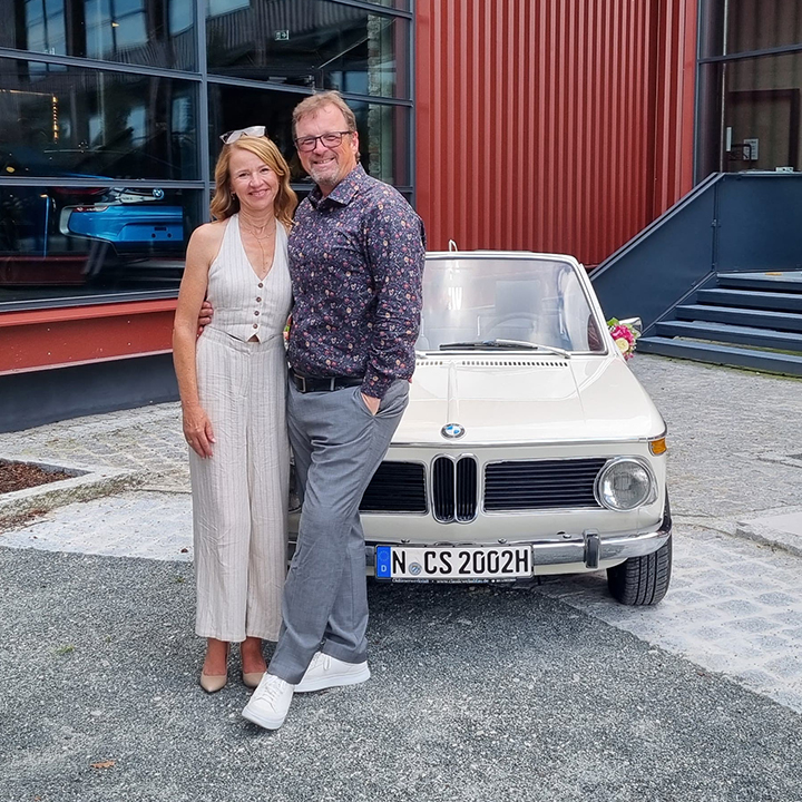 Im Hintergrund sehen wir einen BMW 3er der 1. Generation, vor ihm stehen Christian Silberhorn und eine Frau.
