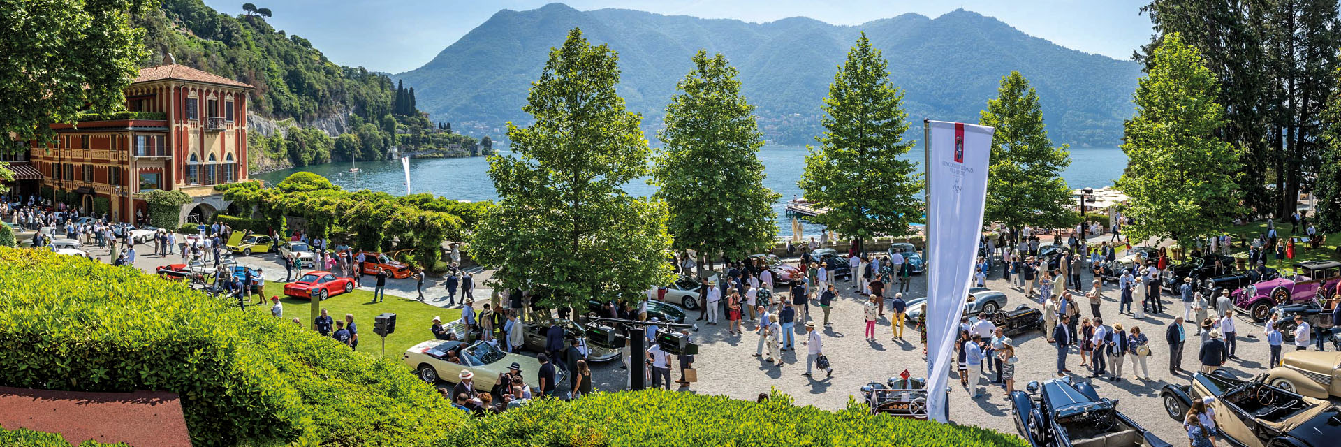 Menschenmenge auf einem Platz mit mehreren geparkten Oldtimern vor einem See und Bergen im Hintergrund.