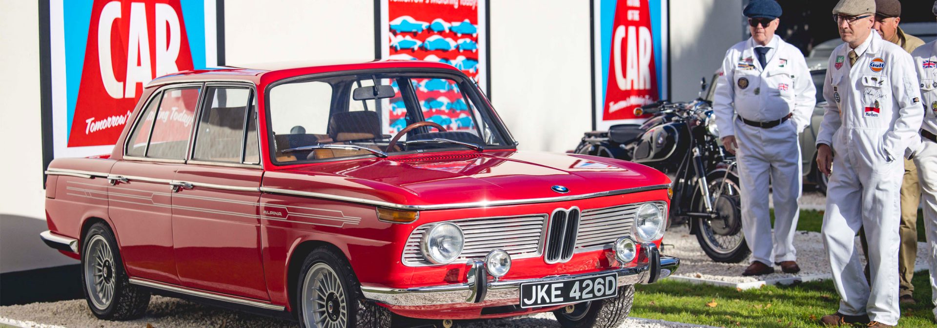 Roter Oldtimer-BMW mit britischem Kennzeichen vor einer Wand mit roten und blauen Werbetafeln, daneben zwei Männer in weißen Overalls und Mützen, im Hintergrund ein Motorrad.