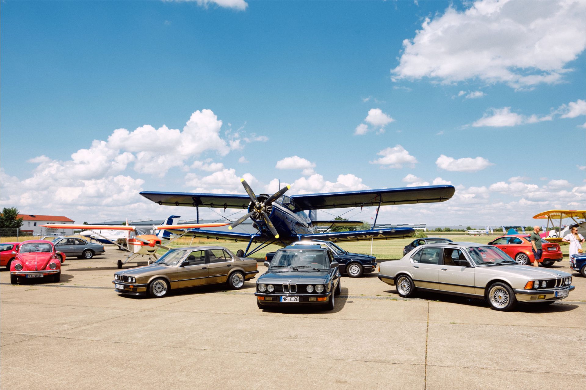 An einem sonnigen Tag sehen wir mehrere BMW Klassiker Automobile vor einem Doppeldecker Flugzeug mit BMW Motor.