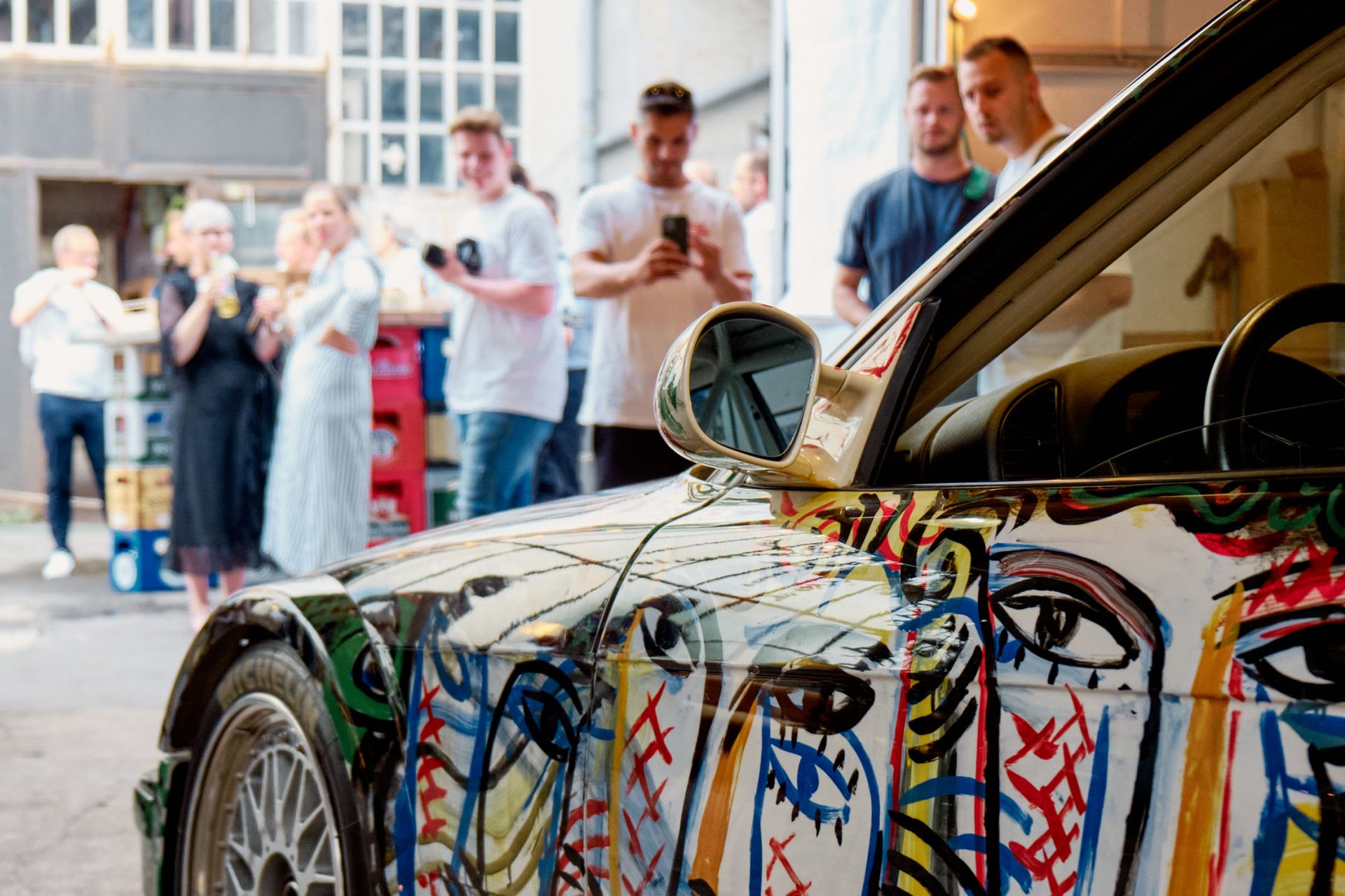 Blick über einen BMW Art Car auf eine Besuchergruppe die teilweise das Fahrzeug fotografiert.