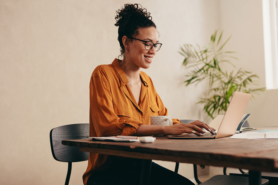 Eine Frau mit Brille sitzt an einem Tisch und beschäftigt sich mit ihrem Laptop.