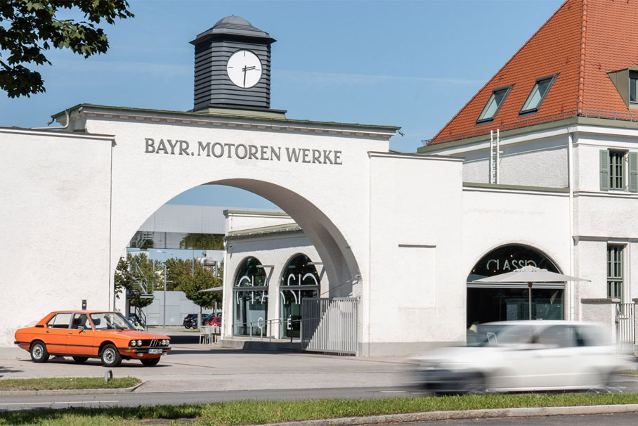 Ein orangener historischer BMW vor dem BMW Group Classic Eingangstor an einem schönen, sonnigen Tag.