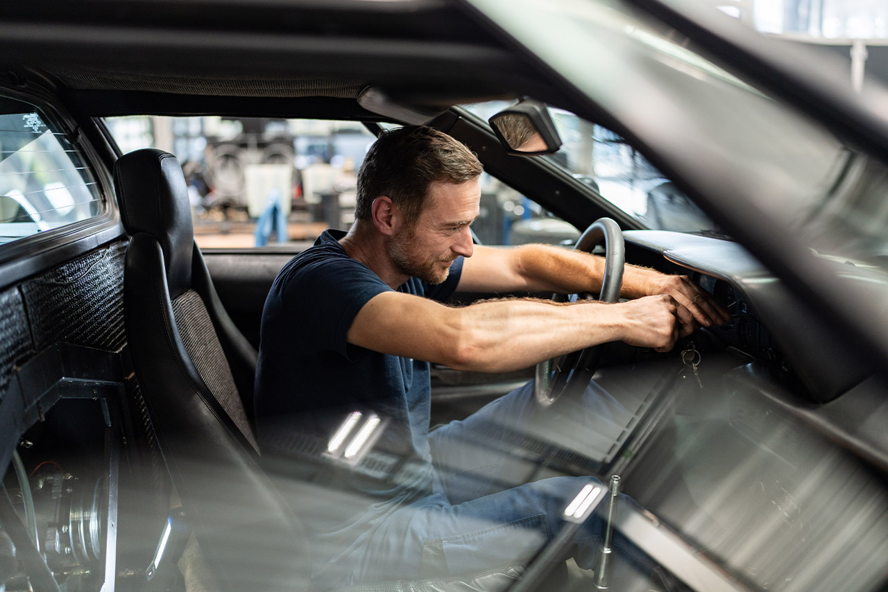 Ein Mann arbeitet an einem BMW Classic Modell in einer Garage, umgeben von Werkzeugen und Autoteilen.
