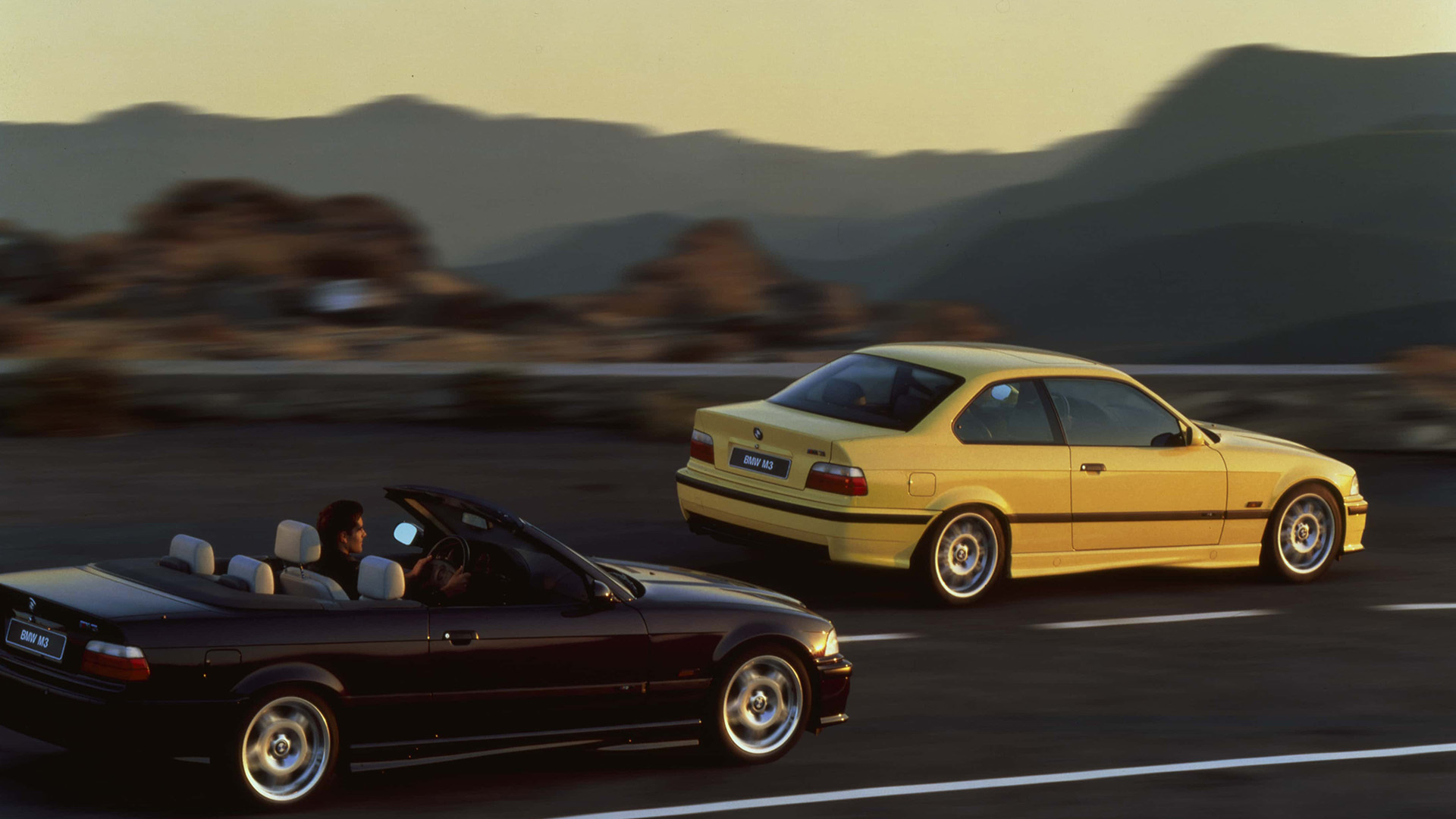 Ein schwarzer und ein gelber 3er BMW der dritten Generation auf einer Landstraße