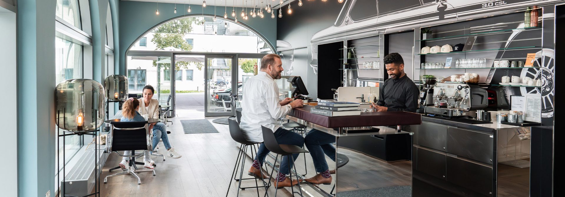 Blick ins BMW Classic Café mit Gästen an der Bar und an Tischen und einem Barista hinter der Theke.