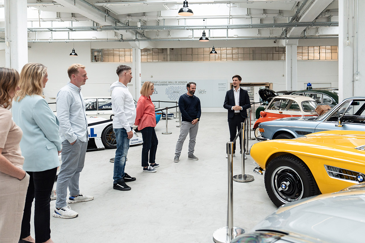 Eine Besuchergruppe hört in den Ausstellungsräumen von BMW Classic aufmerksam einem Museums-Scout zu.