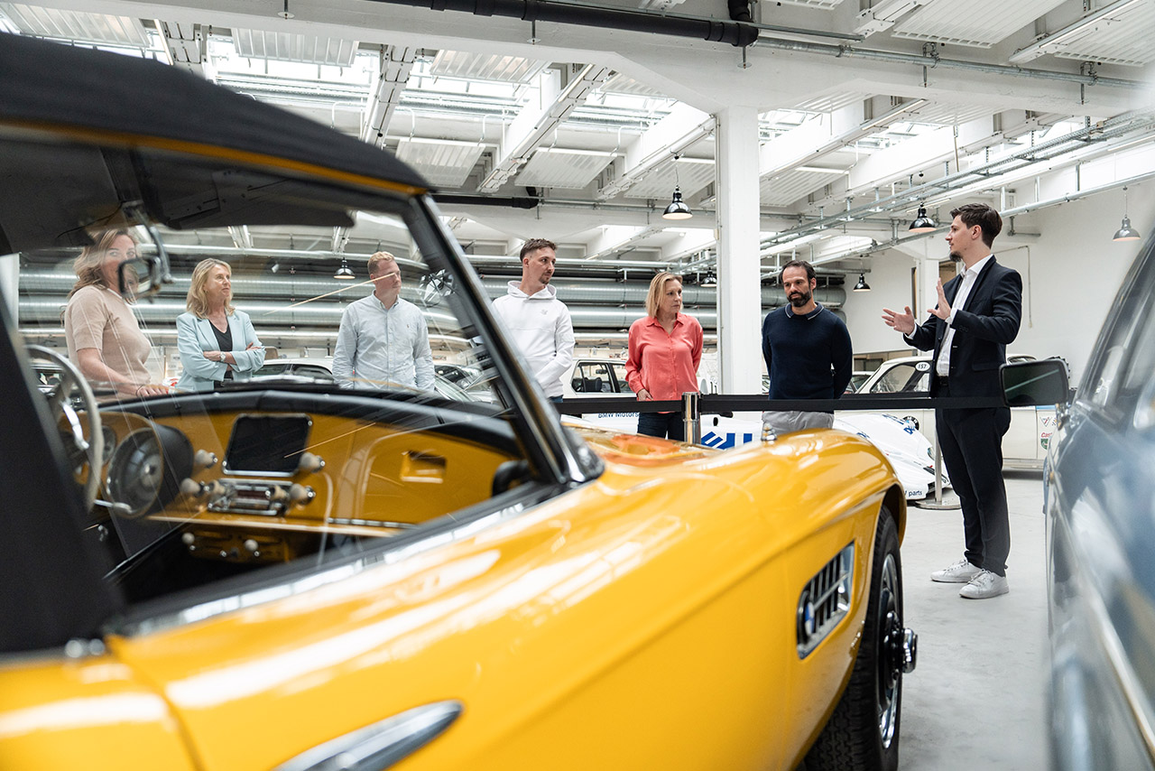 Blick an einem leuchtend gelben 507 Cabrio vorbei auf eine Besuchergruppe in den Ausstellungsräumen von BMW Classic, die aufmerksam einem Museums-Scout lauscht.