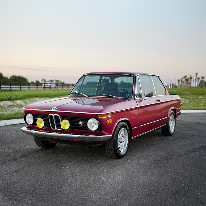 We see a burgundy BMW 2002 from 1974 with yellow auxiliary lights mounted on the bumper on a country road.