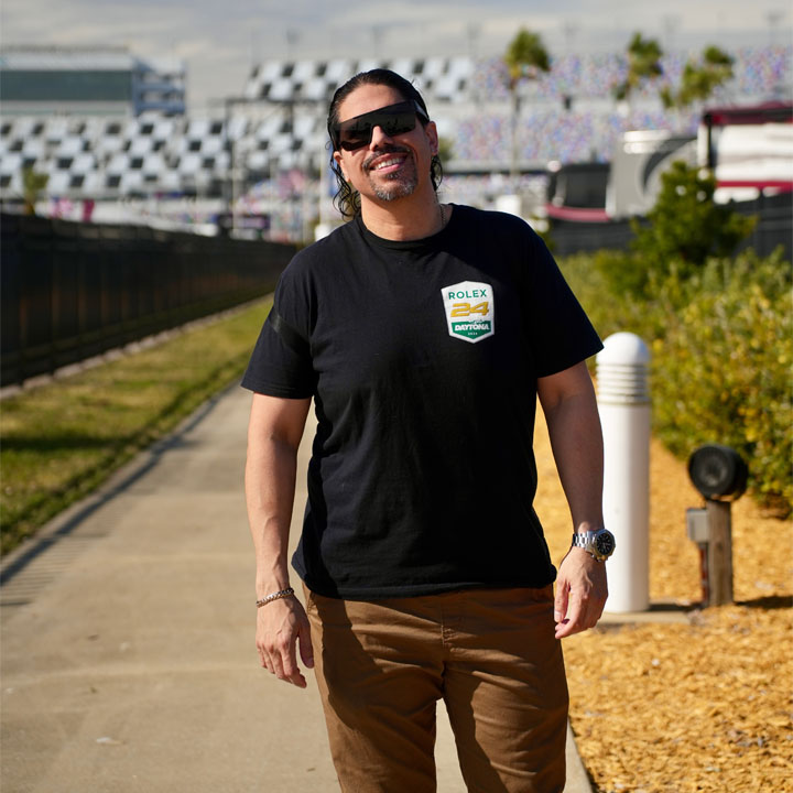 Esteban Valentin, wearing sunglasses, comes towards us on a race track, with black and white checkered track bridges in the background.