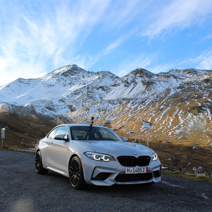 We see a white BMW M2 from the front, parked somewhere on a country road. In the background, snow-capped mountains and a blue sky.