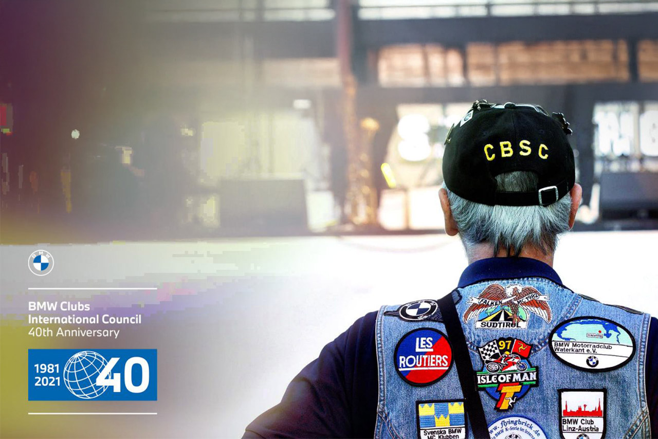 A BMW Classic fan with a cap, grey hair and a denim vest with numerous patches looks away from the viewer towards a building.