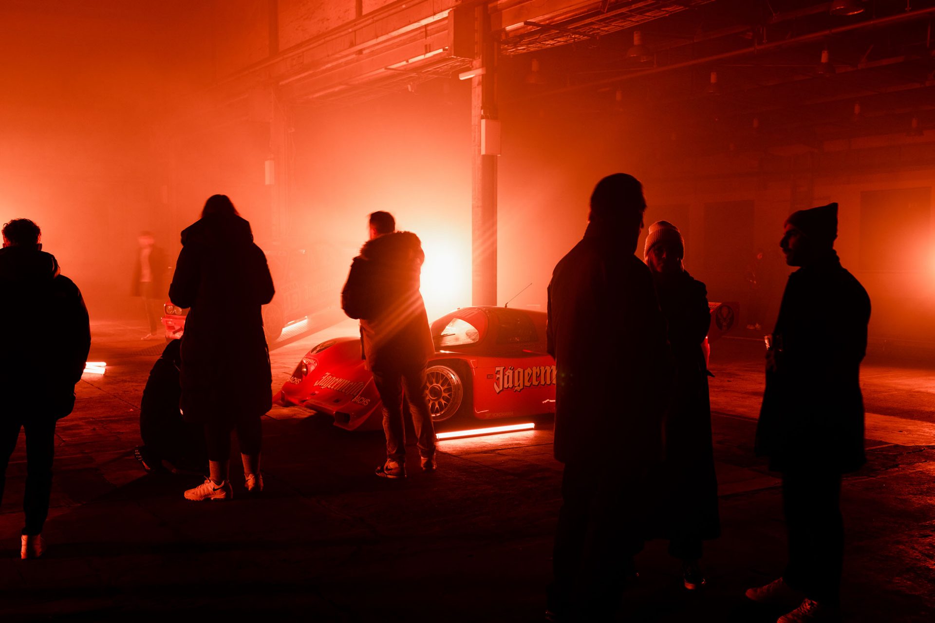 A scene bathed in red light, like those found in clubs, with shadowy figures in the foreground and a BMW racing car in the background.
