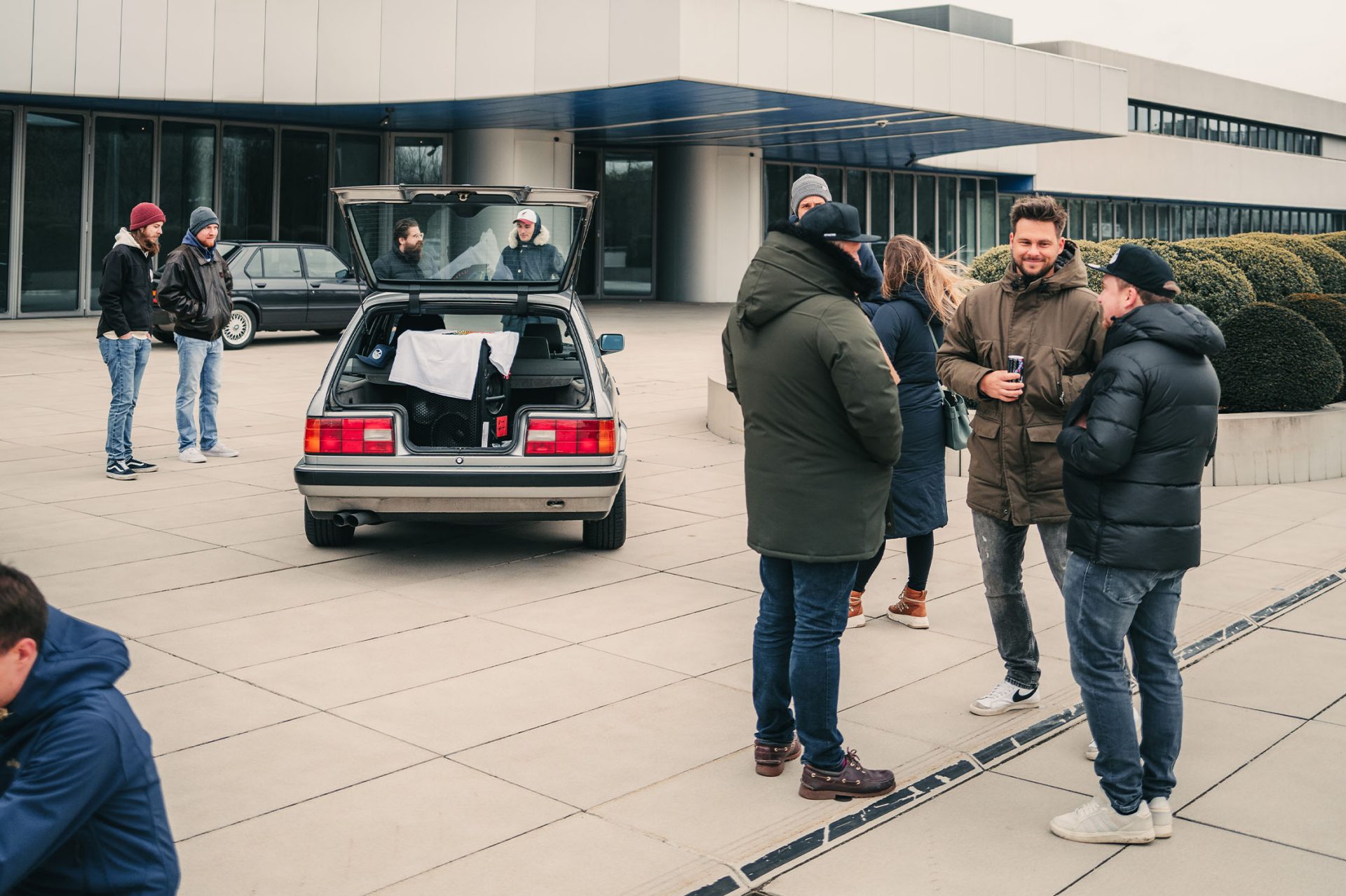 A group of people are standing together talking; next to them is a classic BMW with its tailgate open.