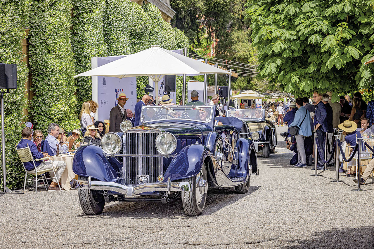 A dark blue convertible from the 1930s drives along a road while people stand on the side of the road and watch.