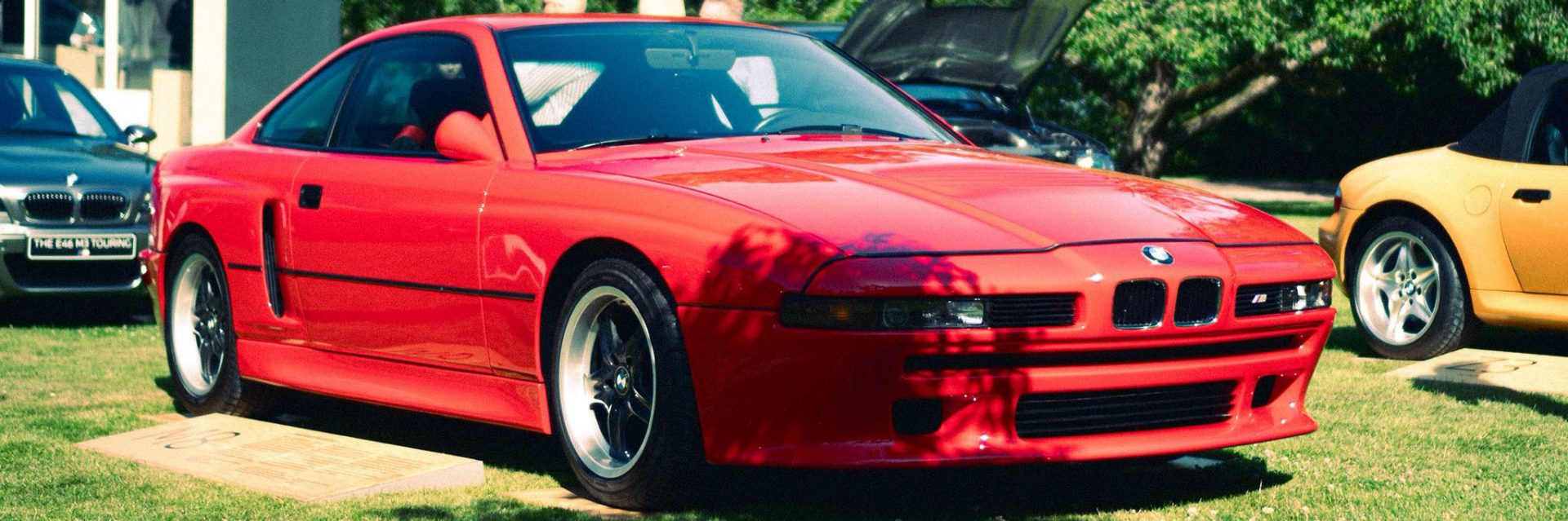 A red BMW sports car from the 1990s in a meadow, with other vehicles in the background.