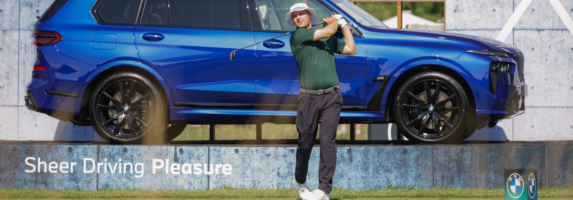 A golfer wearing green clothing and a hat swings a club in front of a blue BMW SUV on a golf course with a billboard.