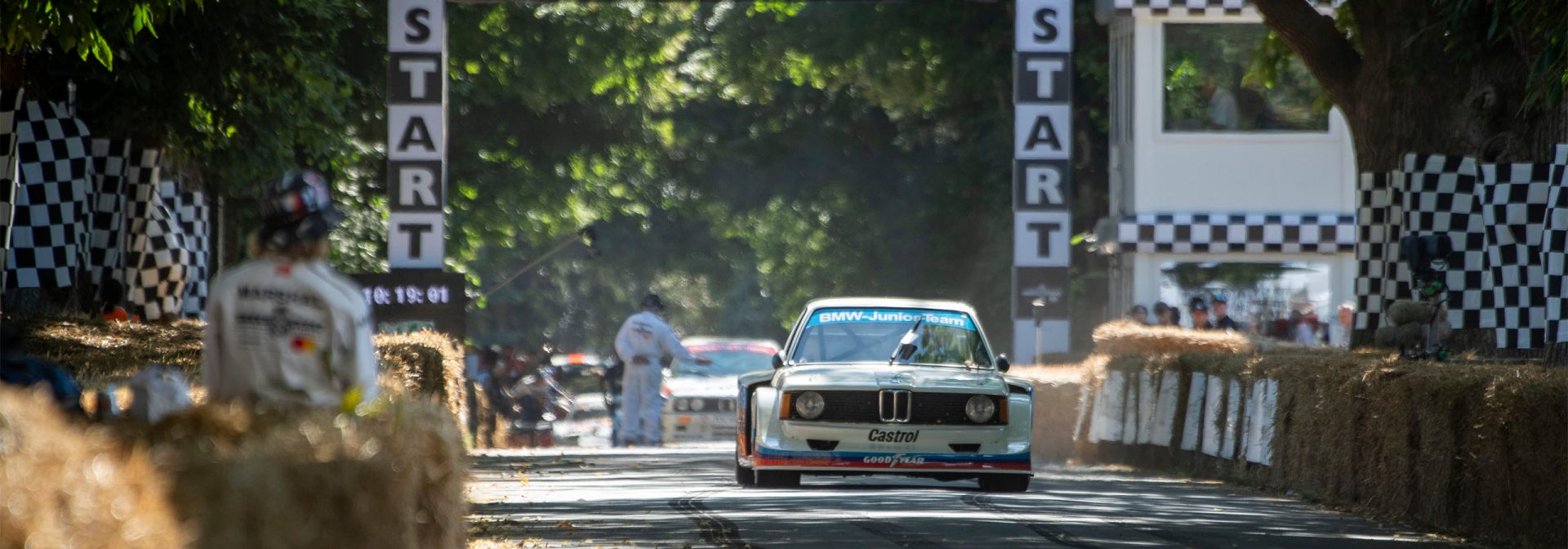 A race car drives along a track lined with bales of straw, passing under a starting banner with a black-and-white checkered pattern.