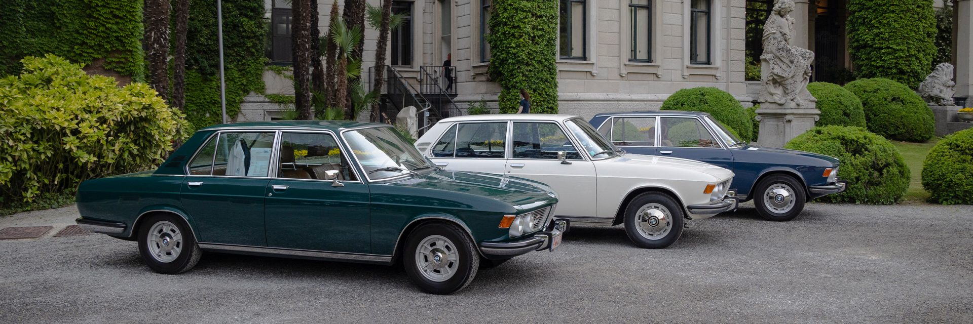 Three classic BMW cars parked in front of a magnificent mansion with elegant architecture.
