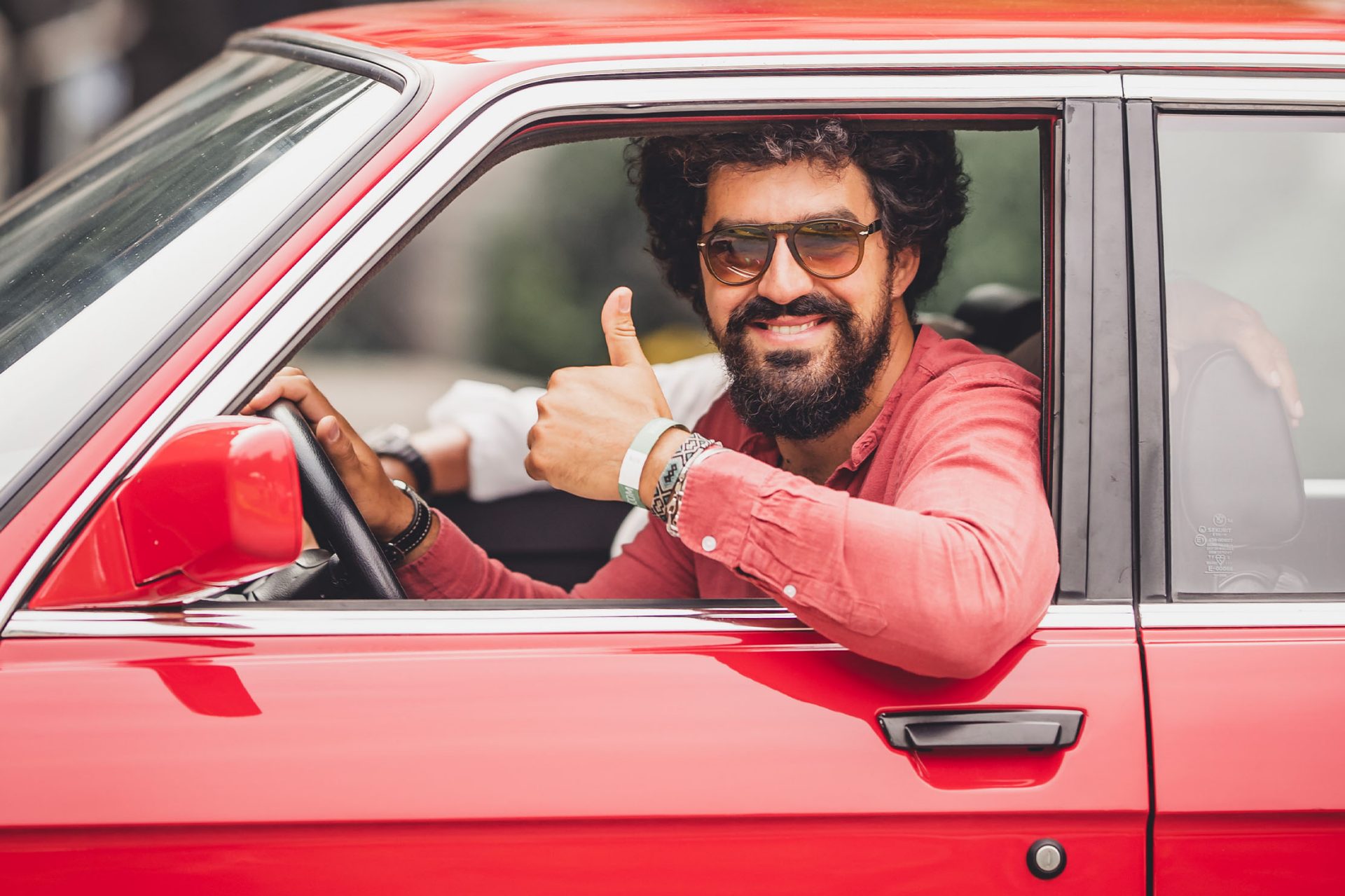 A BMW driver in a red classic BMW greets the viewer with a thumbs up.