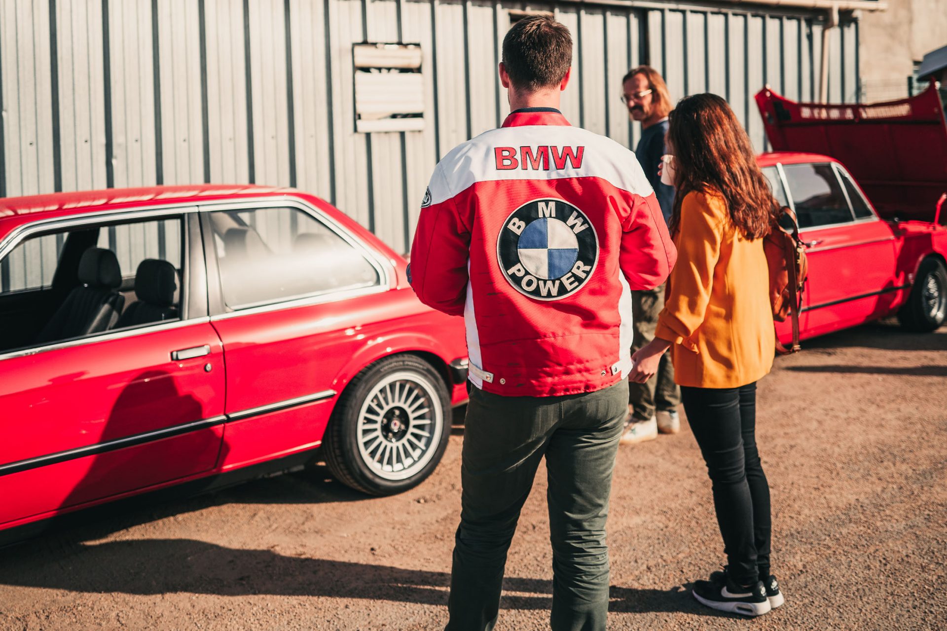 Several BMW fans, one wearing a jacket with a large BMW logo on the back, are standing in front of a red BMW classic.
