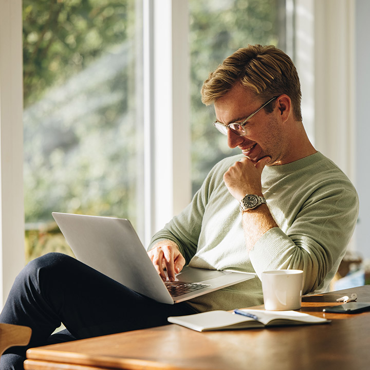 We see a young man, relaxed and smiling as he looks at his laptop in his apartment.