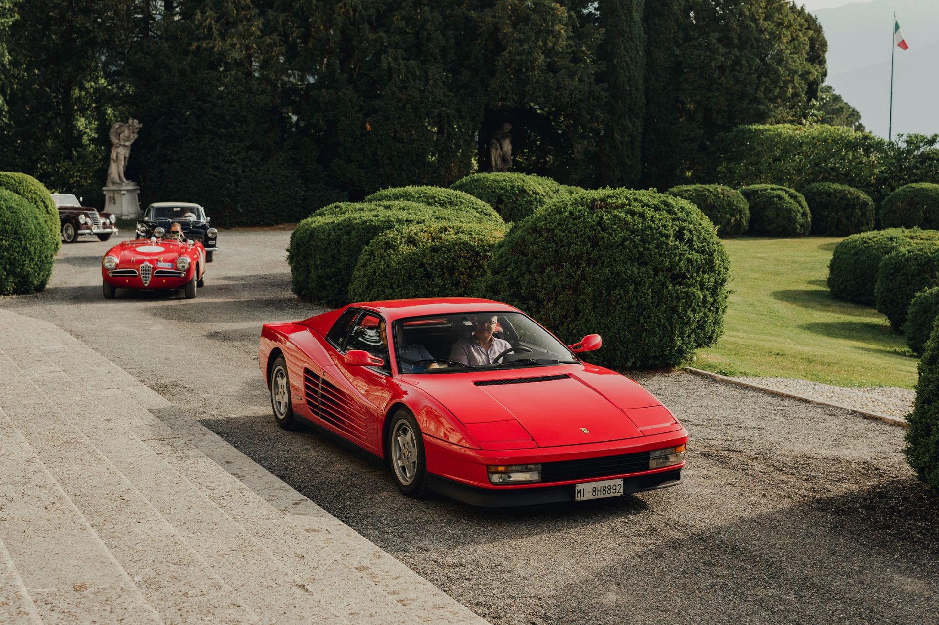 View of the Villa d'Este park through which two red classic cars from Maranello are driving.