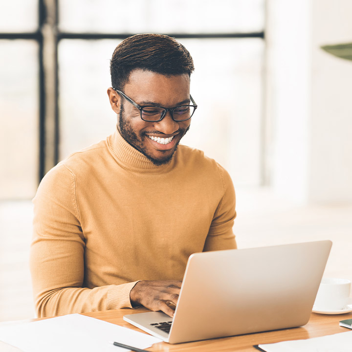 A man with a smile wears a yellow sweater and operates a laptop.