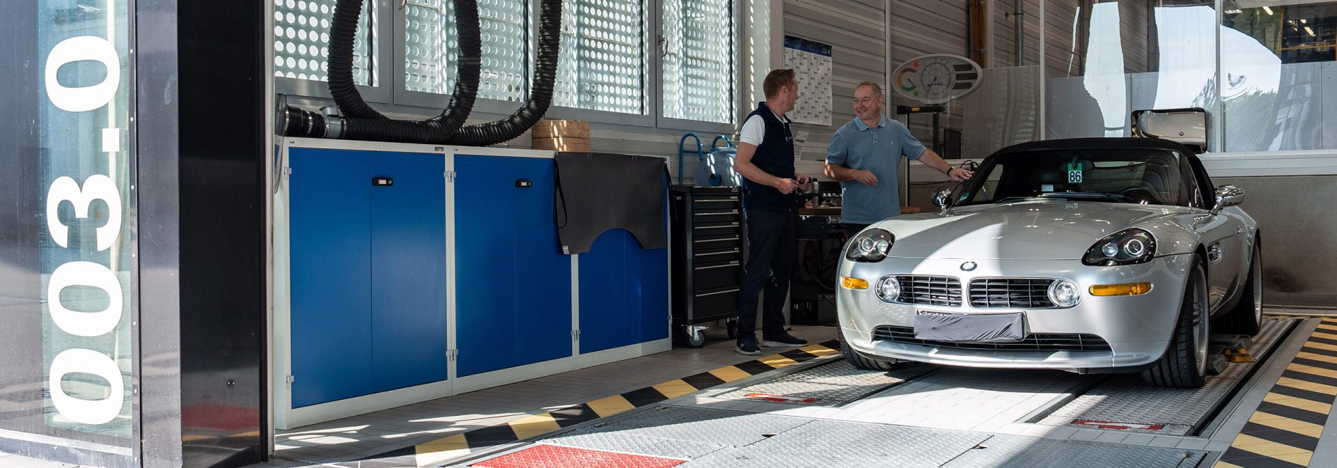 Two men are talking in a BMW Classic garage, surrounded by various tools and a BMW model.